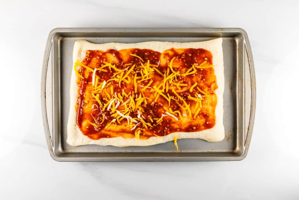 Pizza dough topped with tomato sauce and shredded cheese on a rectangular baking sheet.