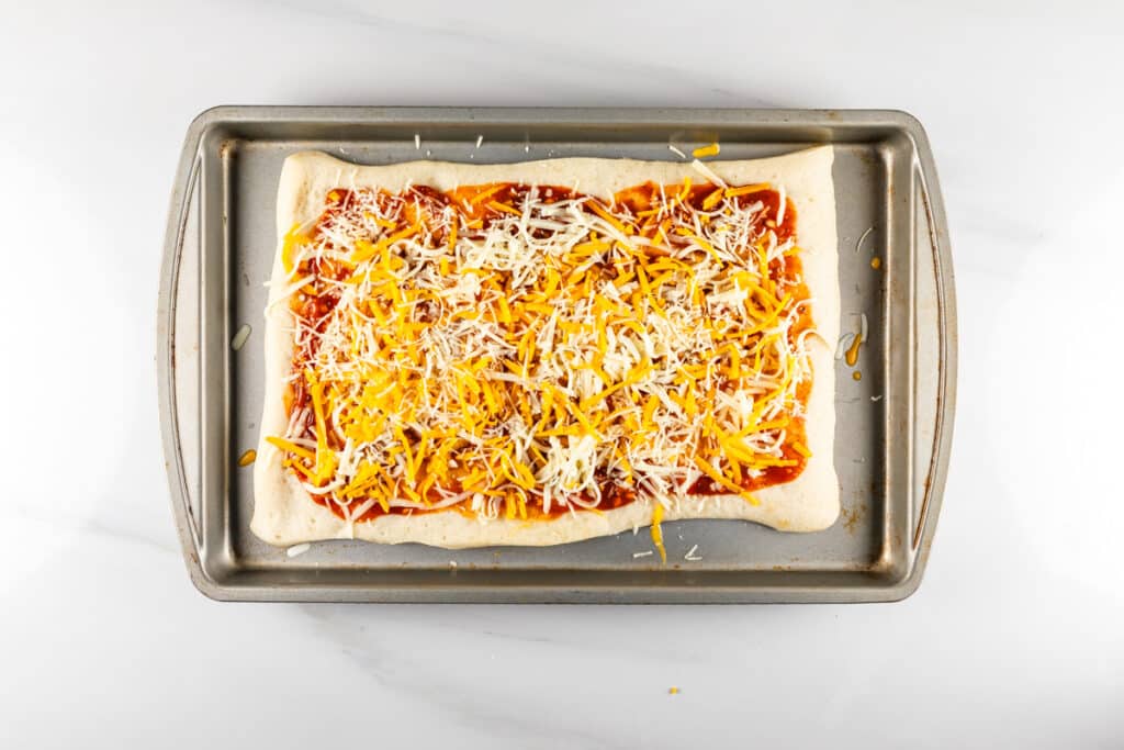 A rectangular pizza dough topped with tomato sauce and a generous layer of shredded cheese, placed on a metal baking sheet. The background is a white marble surface.