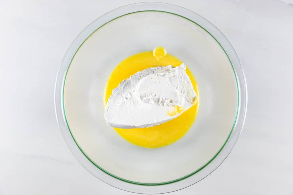 A glass bowl on a marble countertop contains softened cream cheese sitting on melted butter.