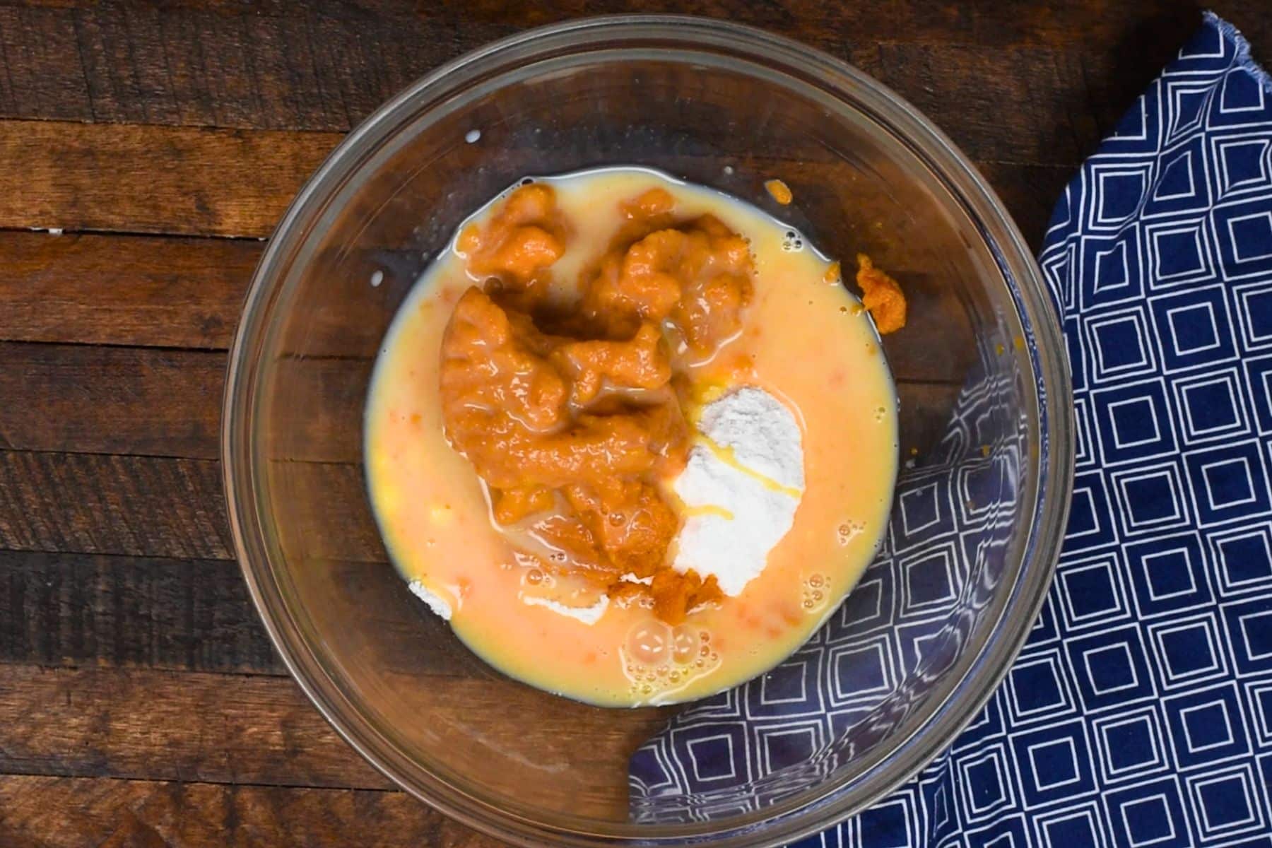 A glass bowl containing a mixture of eggs, pumpkin puree, and flour is on a wooden surface. A blue and white patterned cloth is partially visible on the right.