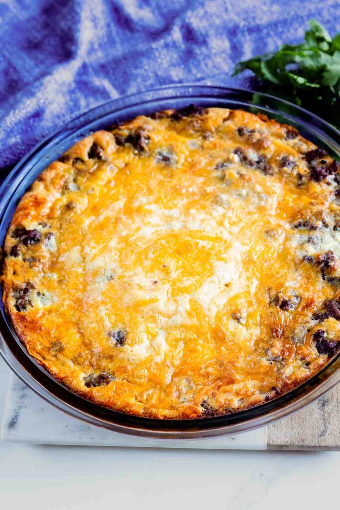 A homemade quiche with a golden brown, cheesy crust is presented in a round baking dish. Bits of browned sausage are visible on the surface. It sits on a white marble board, partially surrounded by sprigs of fresh green parsley.