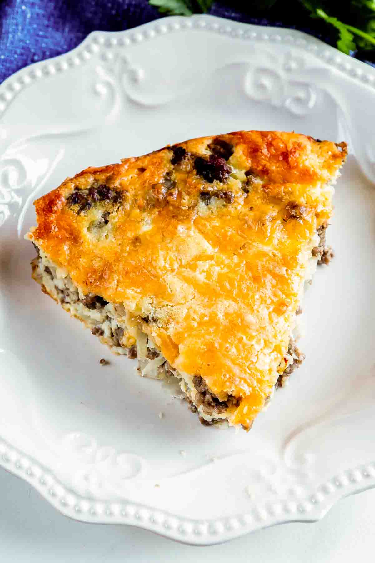 A slice of savory breakfast casserole on a white decorative plate, featuring a golden, melted cheese topping with visible layers of sausage and eggs. The plate sits on a dark blue cloth surface.