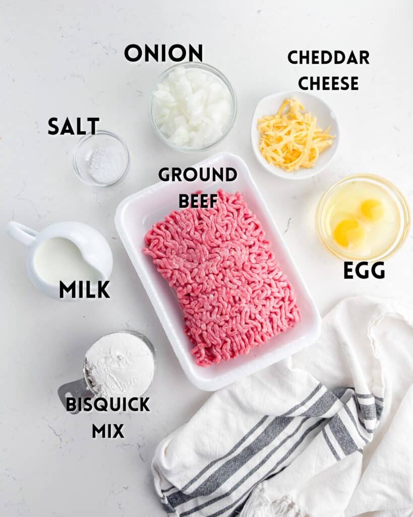 Top-down view of ingredients for a recipe on a white surface: ground beef, chopped onion, cheddar cheese, eggs, milk, Bisquick mix, and salt. A kitchen towel is partially visible to the right. Labels identify each item.