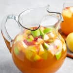 A glass pitcher filled with apple cider punch, containing slices of green and red apples. The background shows a light gray surface with a partial view of an apple and a glass.