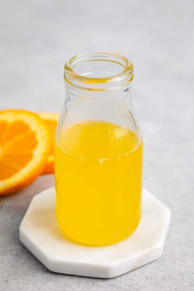 A small glass bottle filled with bright orange juice is placed on a round white coaster. Beside it are two orange slices on a gray surface.