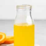 A glass bottle filled with orange juice sits on a round white coaster. In the background, orange slices are placed on a gray surface. The setting is simple and well-lit.