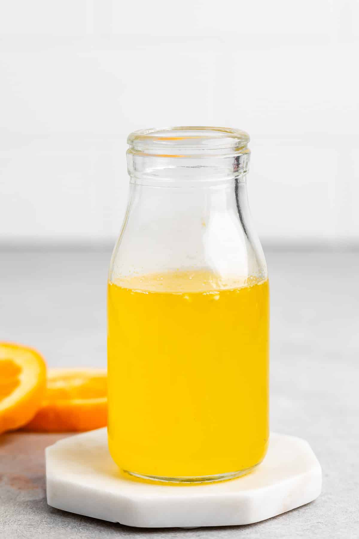 A glass bottle filled with orange juice sits on a round white coaster. In the background, orange slices are placed on a gray surface. The setting is simple and well-lit.