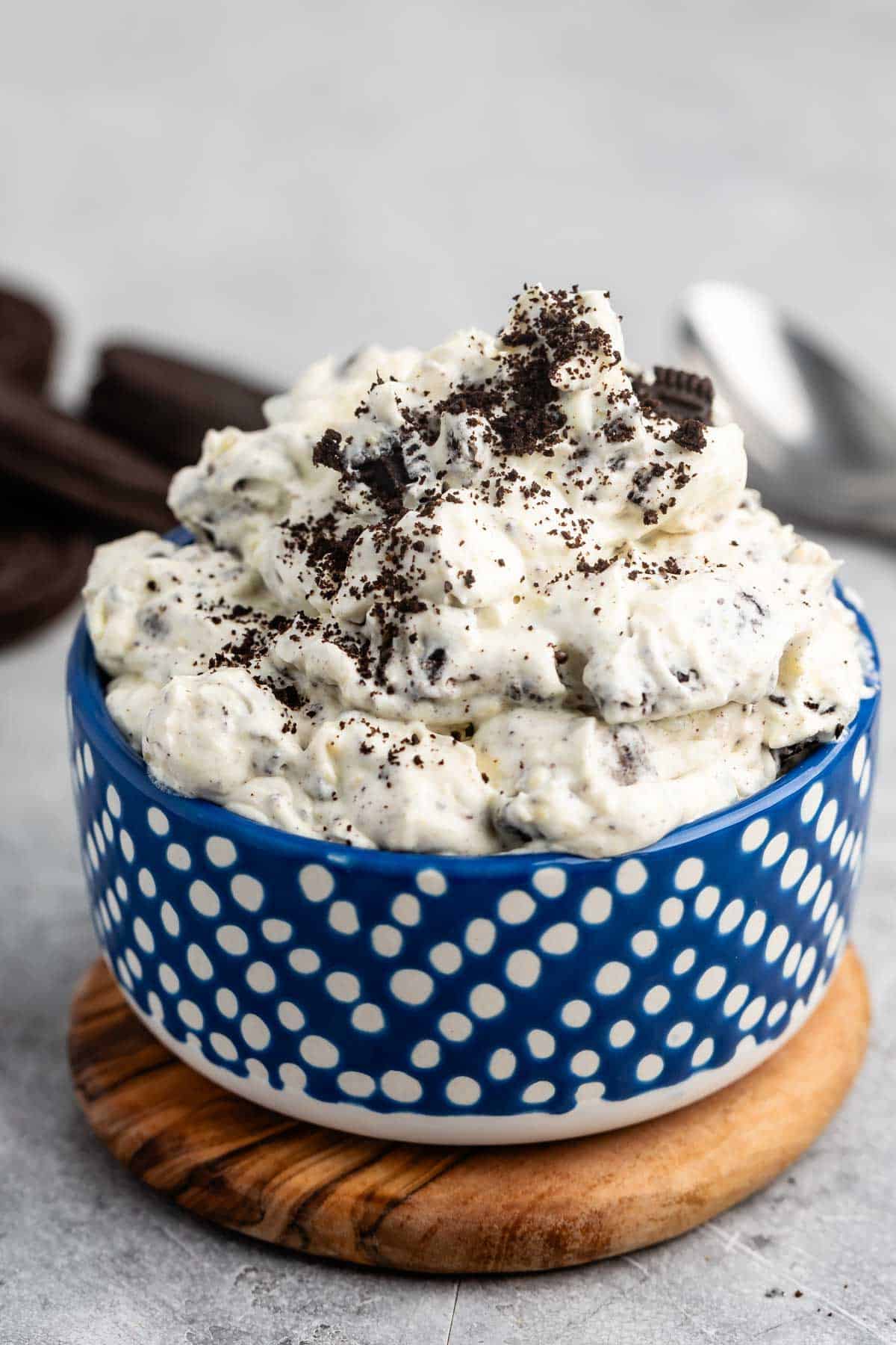 A blue polka dot bowl filled with cookies and cream dip, topped with crushed chocolate cookies, sits on a wooden coaster. A spoon and cookies are blurred in the background on a gray surface.