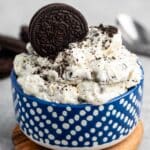 A blue polka-dot bowl filled with a creamy dessert mixed with crushed chocolate cookies, topped with whole and crumbled cookies. The bowl is placed on a wooden coaster on a gray surface. A spoon is visible in the background.