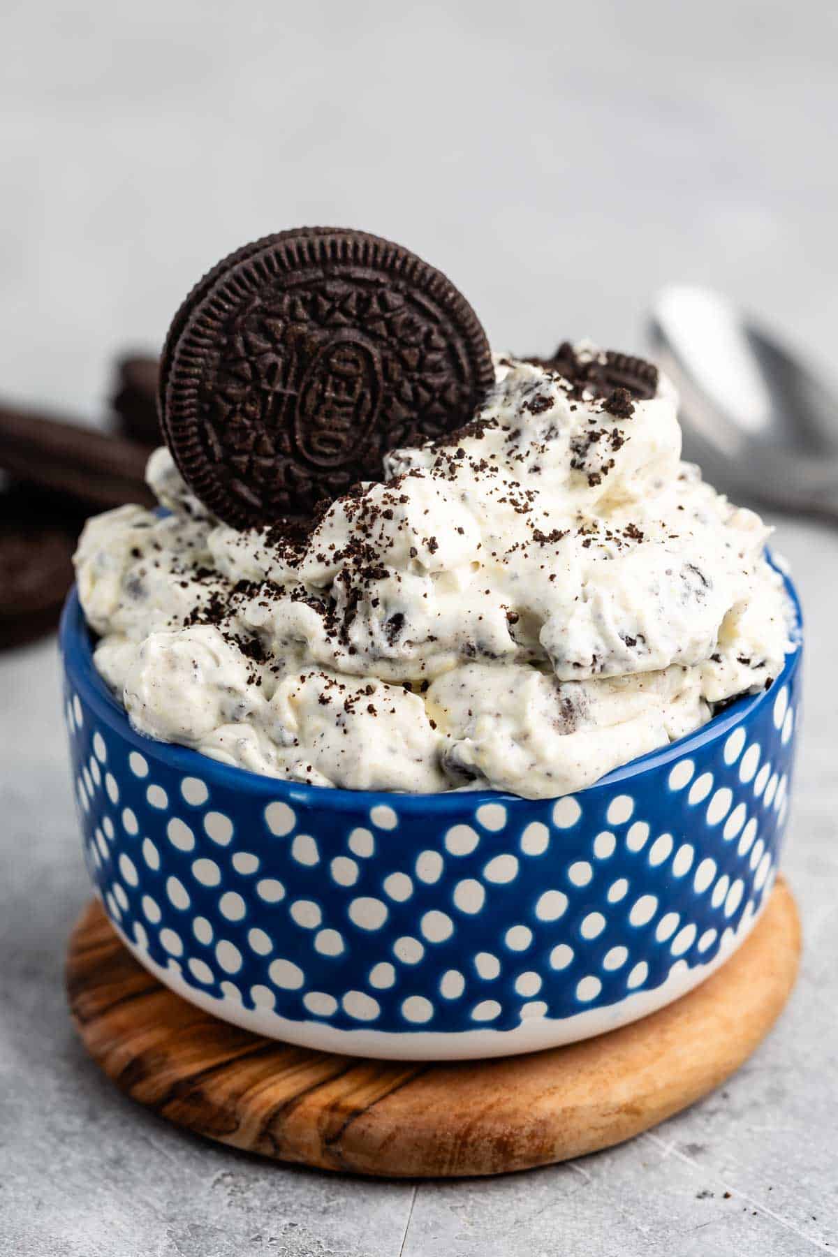 A blue polka-dot bowl filled with a creamy dessert mixed with crushed chocolate cookies, topped with whole and crumbled cookies. The bowl is placed on a wooden coaster on a gray surface. A spoon is visible in the background.