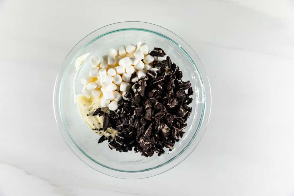 A glass bowl filled with white marshmallows, chocolate sandwich cookie pieces, and a creamy mixture on a white countertop.