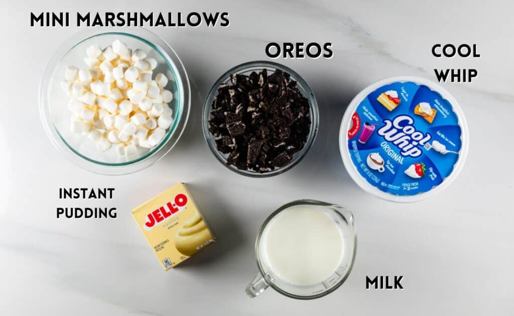 A top-down view of dessert ingredients on a white surface: a bowl of mini marshmallows, crushed Oreos, a tub of Cool Whip, a box of Jello instant pudding, and a glass measuring cup of milk.