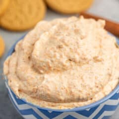 A blue and white chevron-patterned bowl filled with creamy pumpkin dip. In the background, there are round cookies and a cinnamon stick on a gray surface.