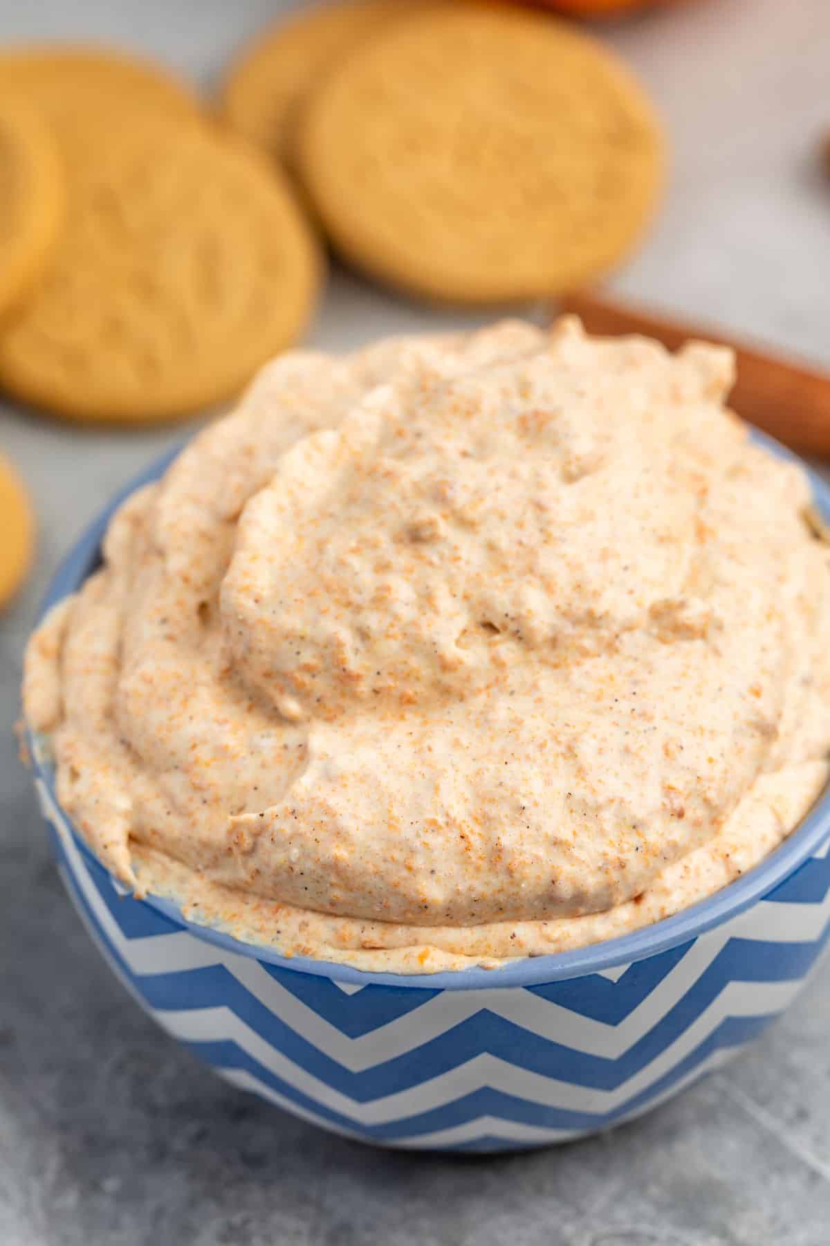 A blue and white chevron-patterned bowl filled with creamy pumpkin dip. In the background, there are round cookies and a cinnamon stick on a gray surface.