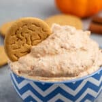 A blue and white zigzag bowl filled with a creamy pumpkin dip, garnished with a ginger cookie. In the background, there is a small pumpkin and more cookies on a gray surface.