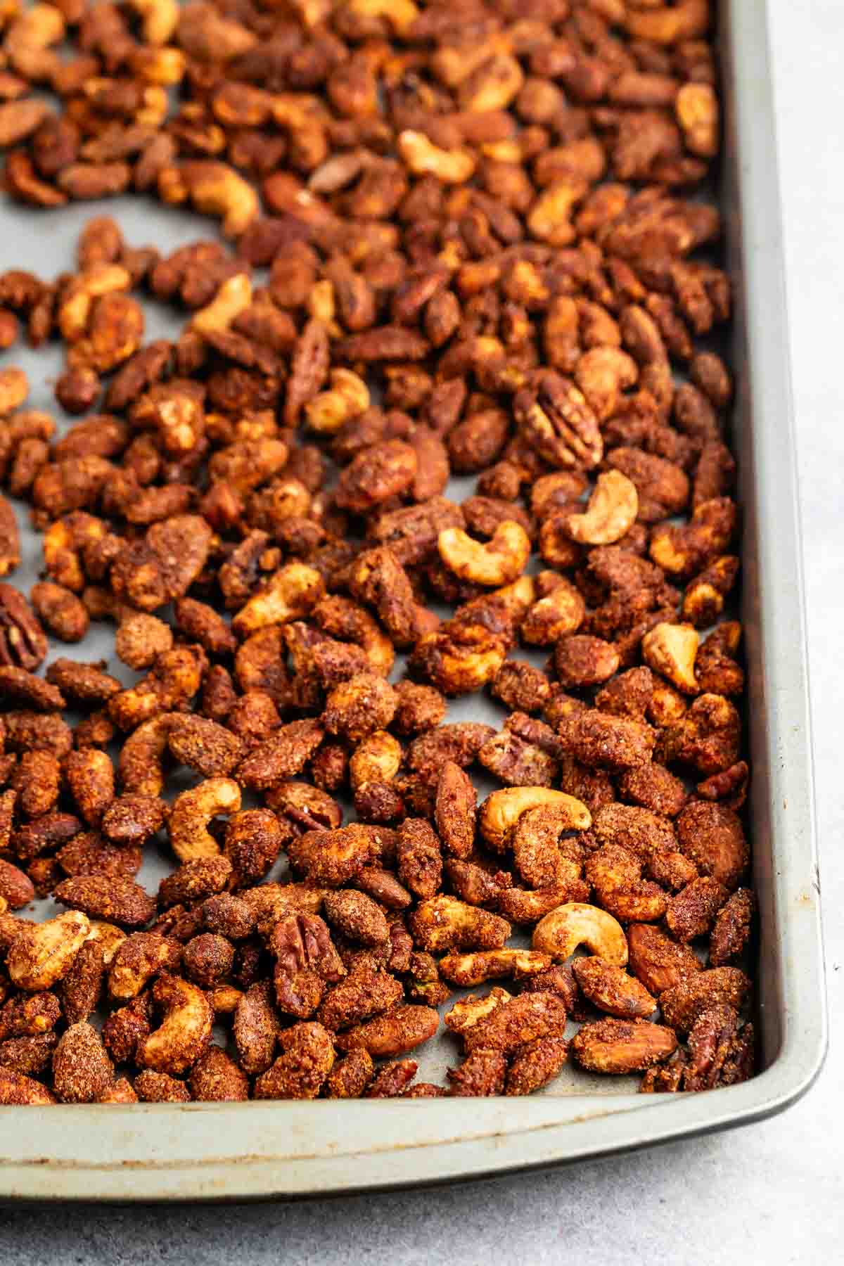 A baking tray filled with a variety of spiced, roasted nuts, including almonds, cashews, and pecans, evenly spread out on a grey surface. The nuts are coated with a brown, spiced seasoning.