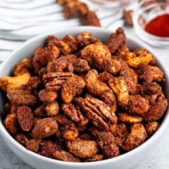 A bowl filled with roasted mixed nuts coated in spices. Two small bowls with powdered spices are in the background on a striped cloth. The nuts appear to include almonds, pecans, and cashews.