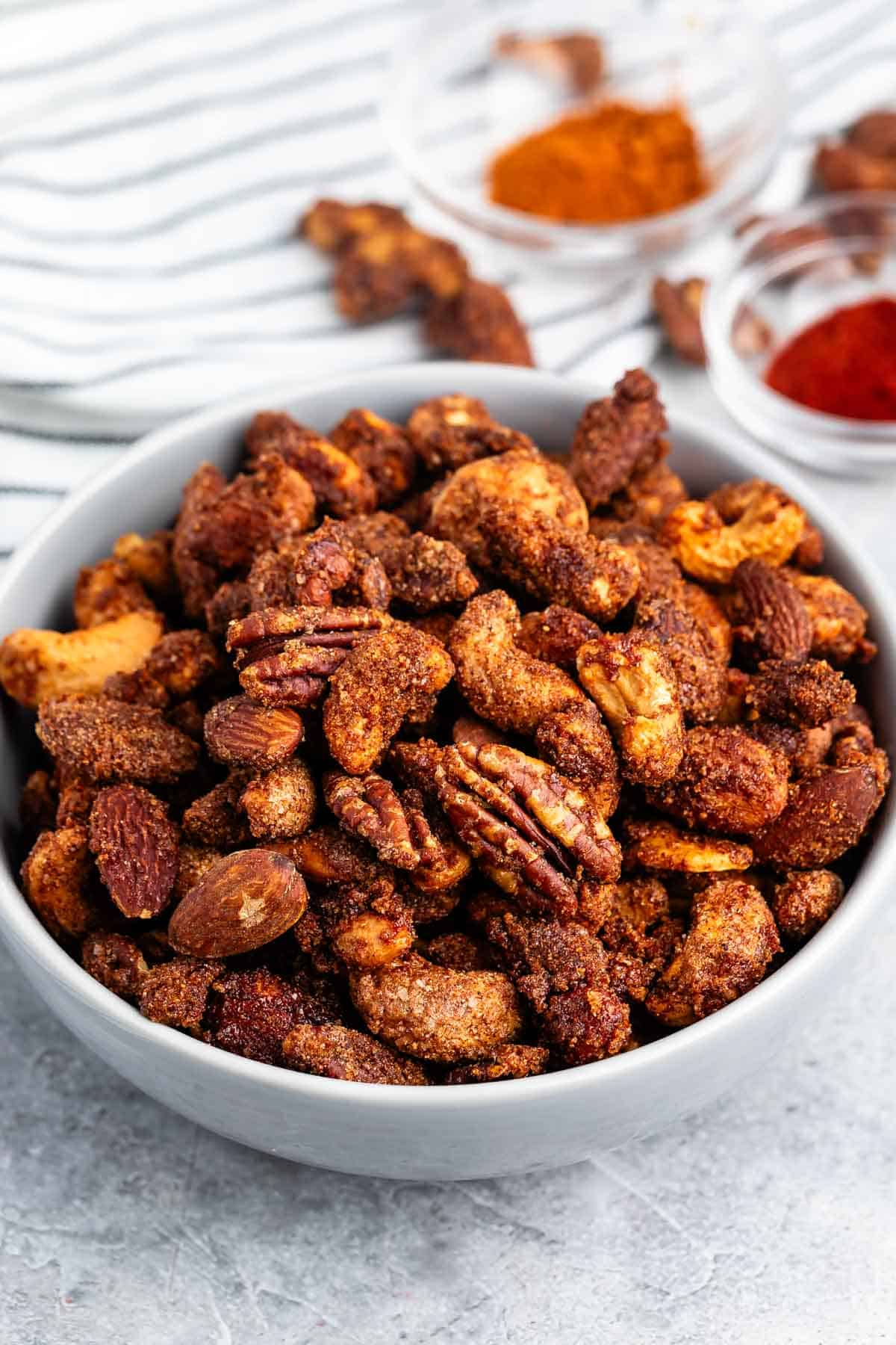 A bowl filled with roasted mixed nuts coated in spices. Two small bowls with powdered spices are in the background on a striped cloth. The nuts appear to include almonds, pecans, and cashews.