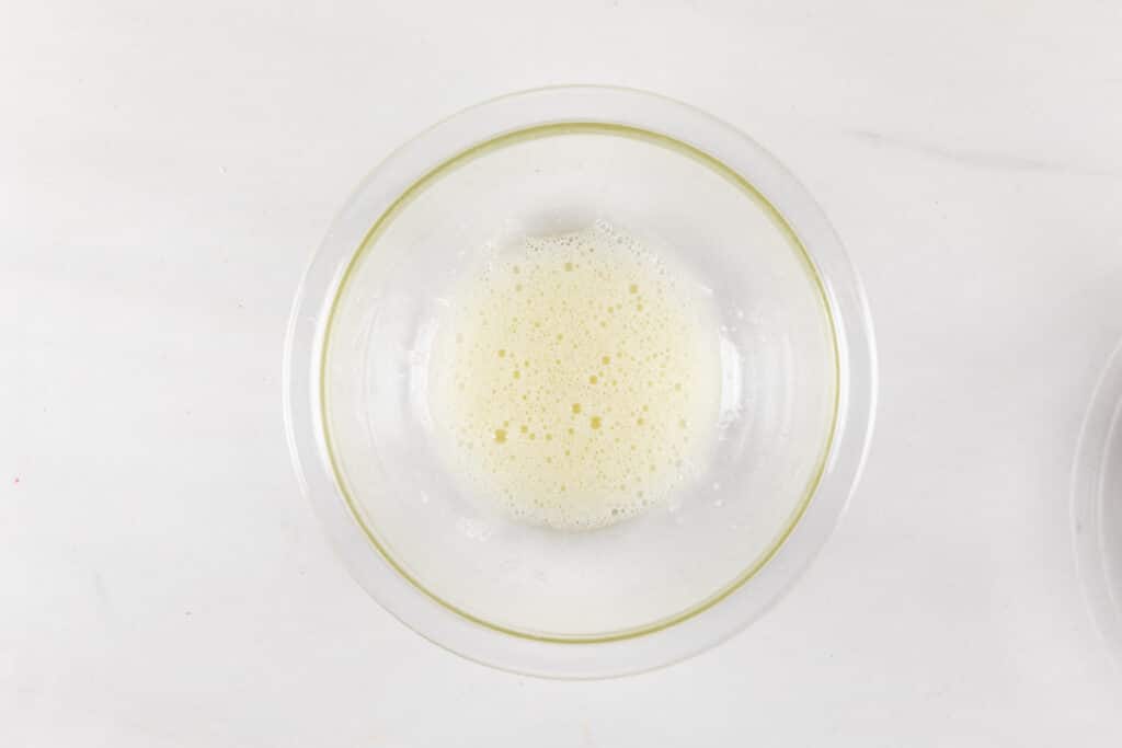 A clear glass bowl containing a frothy mixture on a white surface. The foamy substance appears light and airy.