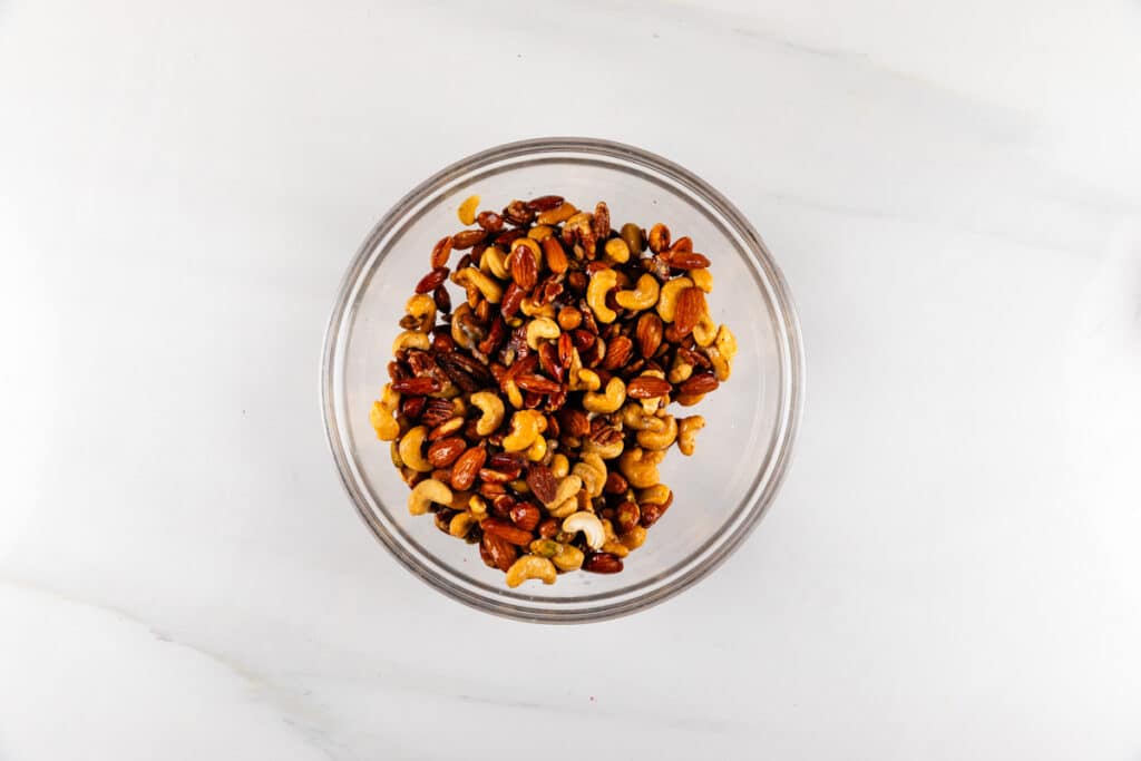 A clear glass bowl filled with an assortment of mixed nuts, including almonds, cashews, and pecans, sits on a light marble surface.