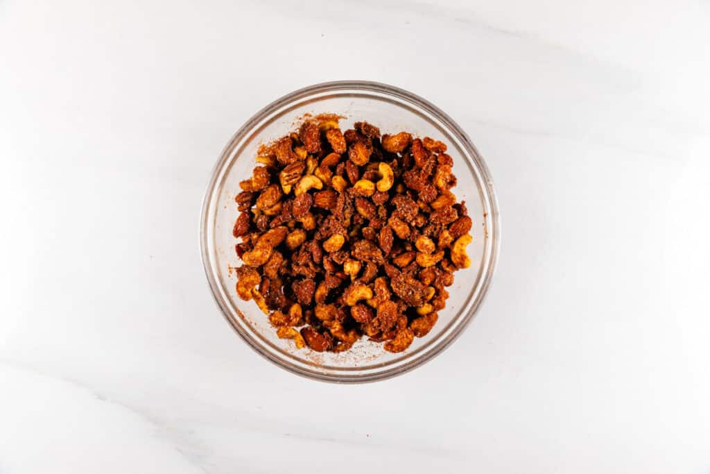 A glass bowl filled with roasted mixed nuts, coated in a brown, spiced seasoning, sits on a white marble surface. The nuts appear crunchy and flavorful.