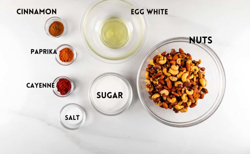 Ingredients for spiced nuts on a white surface: a bowl of mixed nuts, a bowl of egg white, small bowls of sugar, salt, cinnamon, paprika, and cayenne pepper.