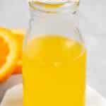 A small glass bottle filled with bright orange liquid sits on a white coaster. An orange slice is visible in the background. Text at the top reads No Alcohol Triple Sec Recipe and easygoodideas.com at the bottom.