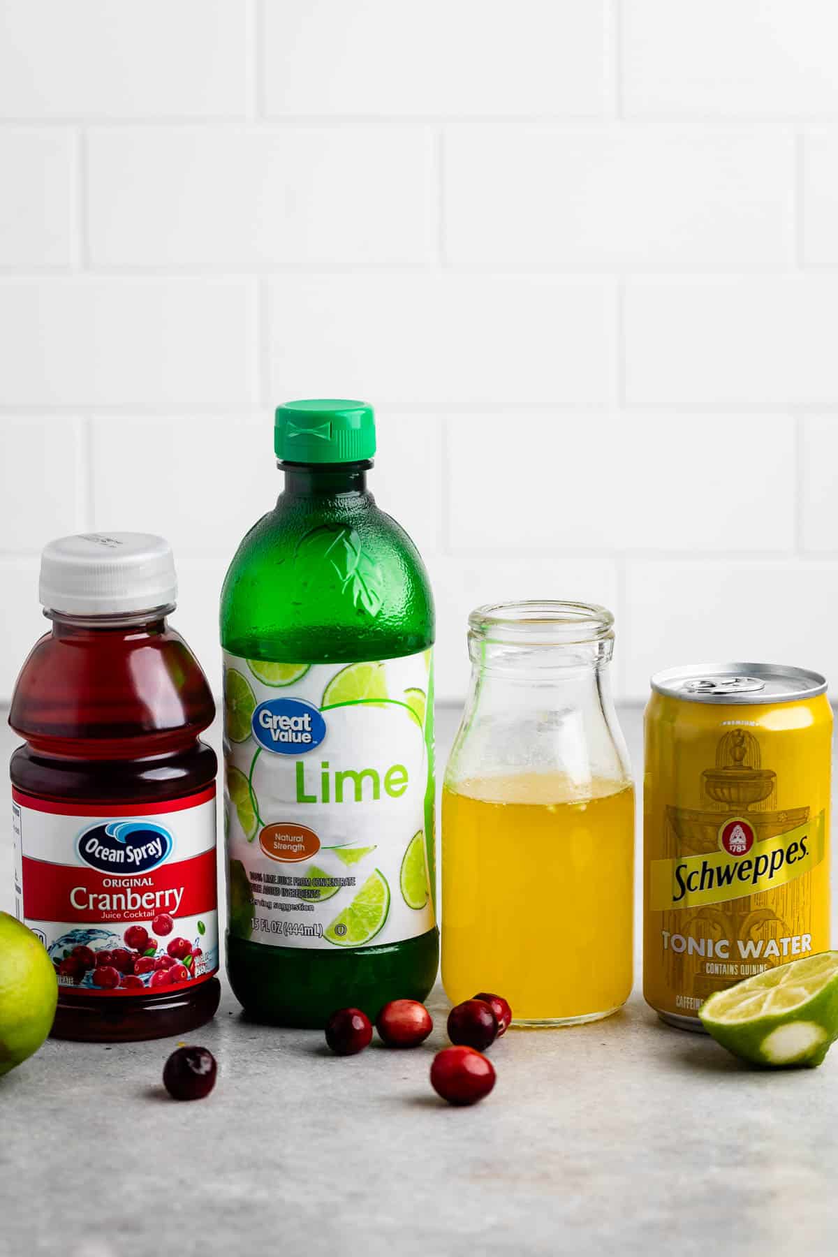 Bottles of cranberry juice and lime juice, a can of tonic water, and a small glass bottle of yellow liquid are arranged on a counter. Additional elements include whole cranberries and lime wedges scattered around.