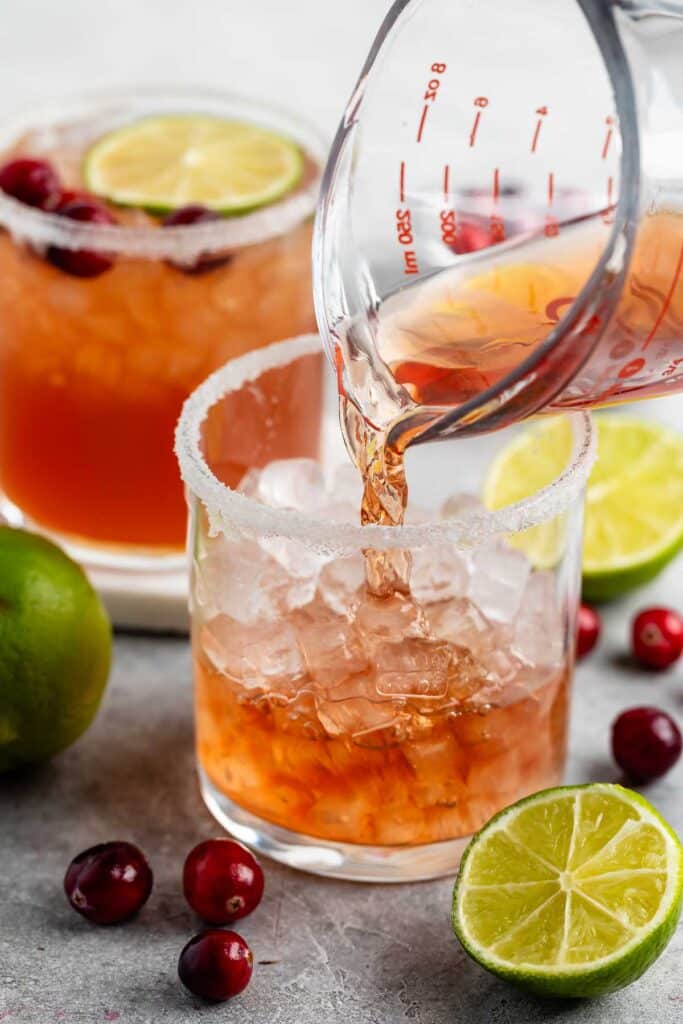 A close-up of a drink being poured from a measuring glass into a sugar-rimmed glass filled with ice. The drink is reddish-brown. Nearby are limes and cranberries. Another glass with a similar drink and garnish is in the background.