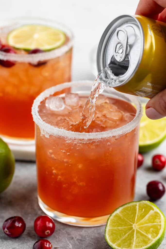 A fizzy drink is being poured from a can into a glass with a salted rim. The drink is light red and contains ice cubes. Surrounding the glass are slices of lime and scattered cranberries; another similar drink is in the background.