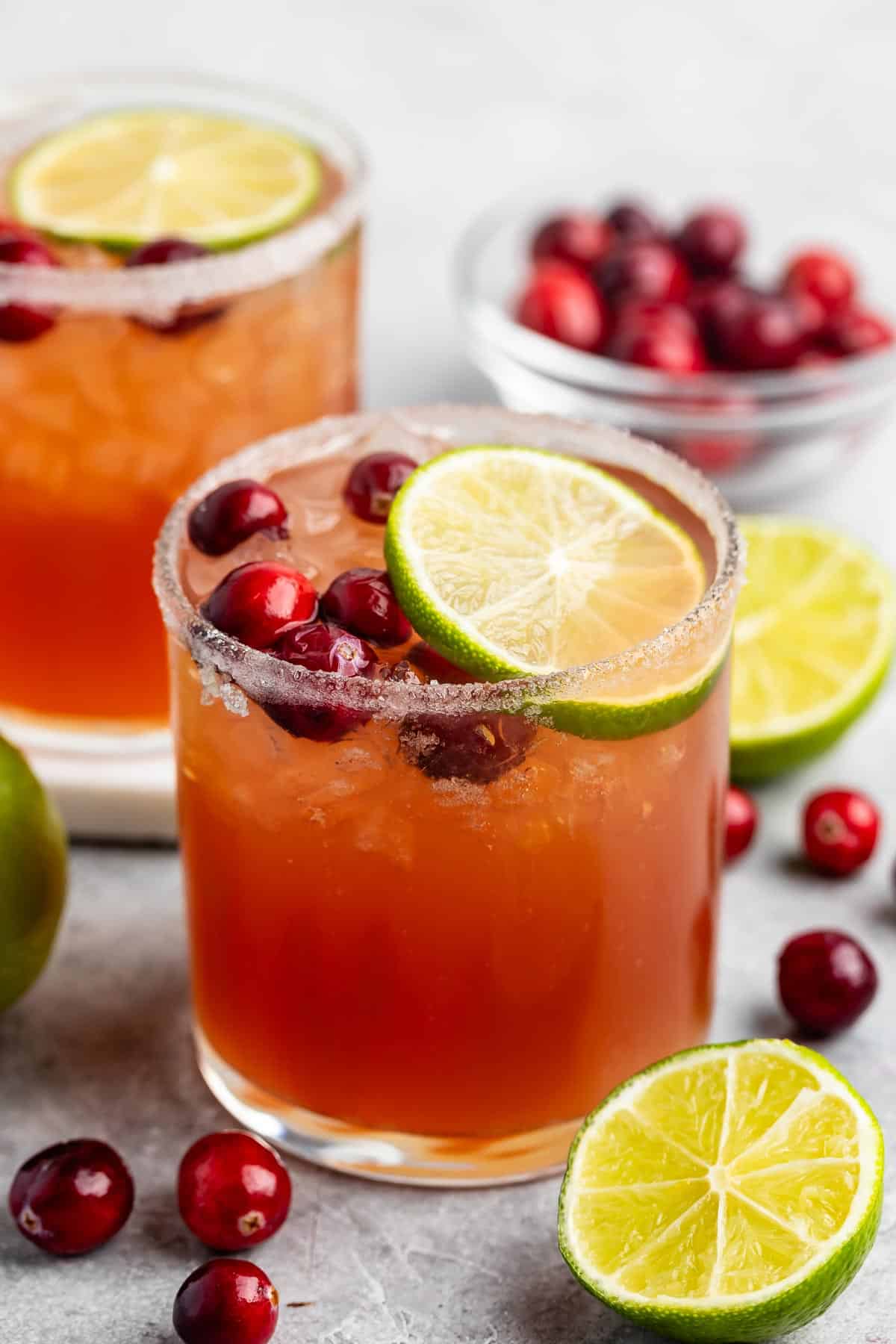 Two glasses of cranberry margaritas with salt-rimmed edges and garnished with lime slices and cranberries. Fresh cranberries and lime halves are scattered around, with a small bowl of cranberries in the background.