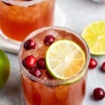 Two glasses of iced cranberry cocktail garnished with lime slices and cranberries, rimmed with sugar. Fresh cranberries and lime halves are scattered around on a gray surface.