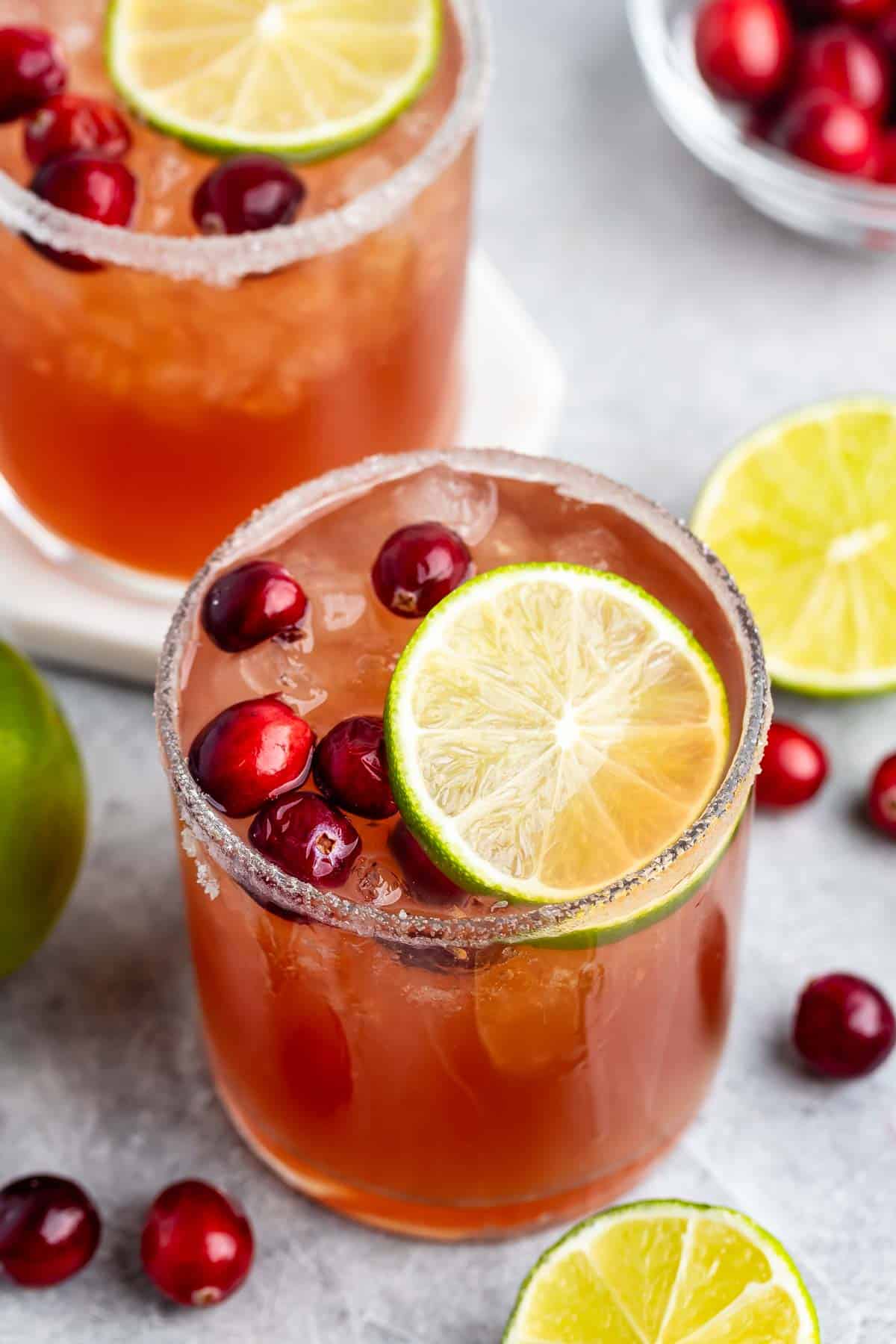 Two glasses of iced cranberry cocktail garnished with lime slices and cranberries, rimmed with sugar. Fresh cranberries and lime halves are scattered around on a gray surface.