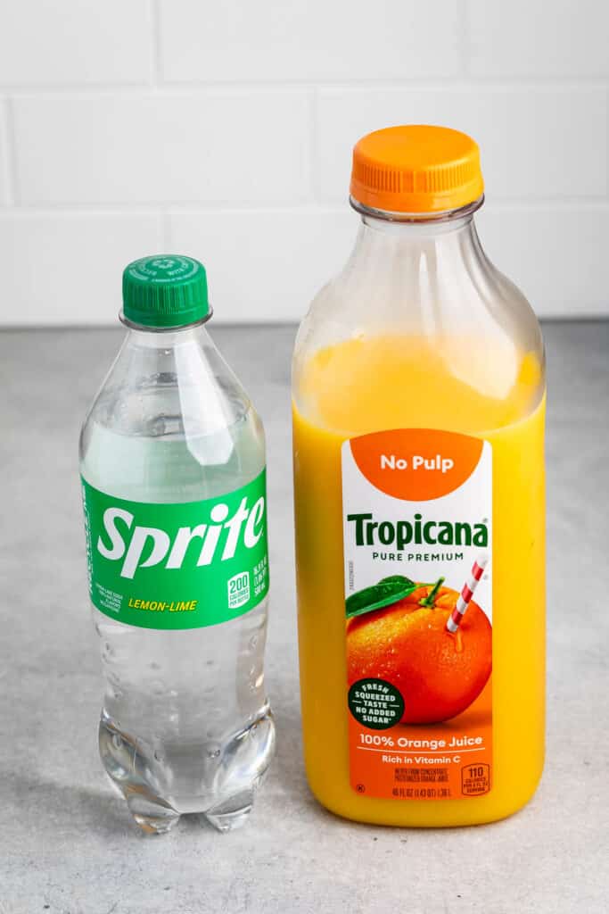 A bottle of Sprite lemon-lime soda next to a Tropicana Pure Premium orange juice bottle on a gray surface. The orange juice label indicates No Pulp and 100% Orange Juice.