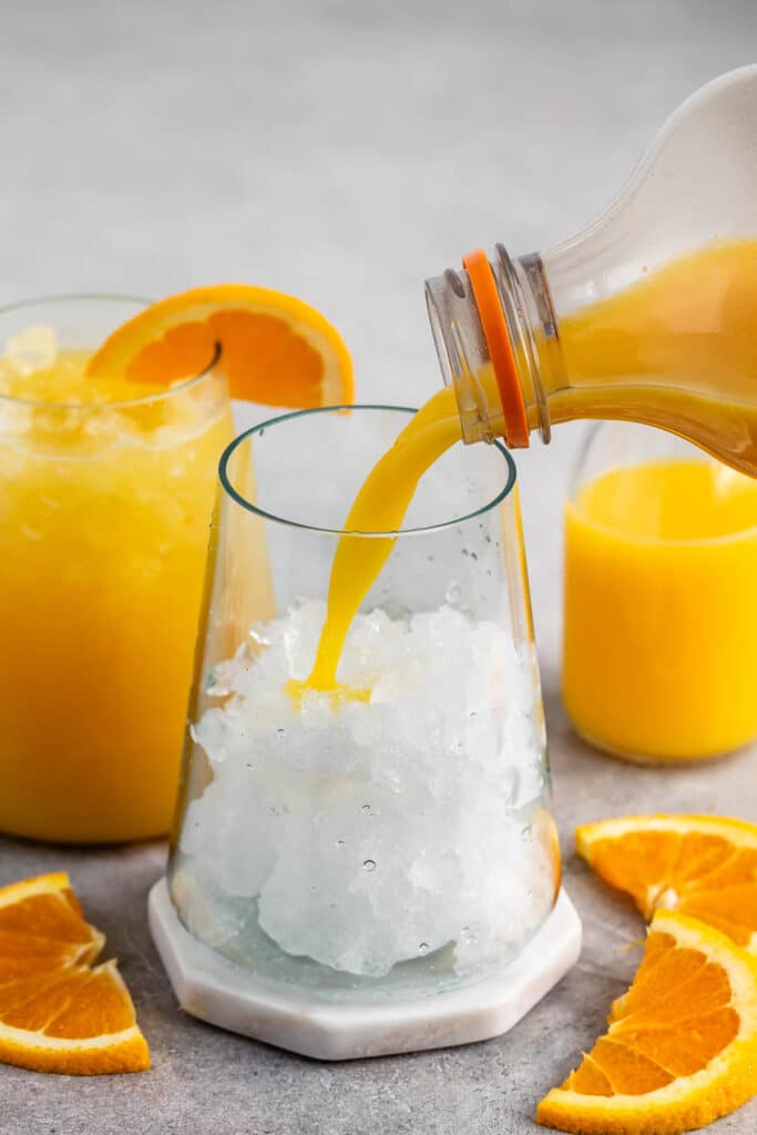 A glass is being filled with orange juice over crushed ice. Nearby, a bottle of juice and a glass with juice and an orange slice garnish are visible. Slices of fresh orange are placed on the table.