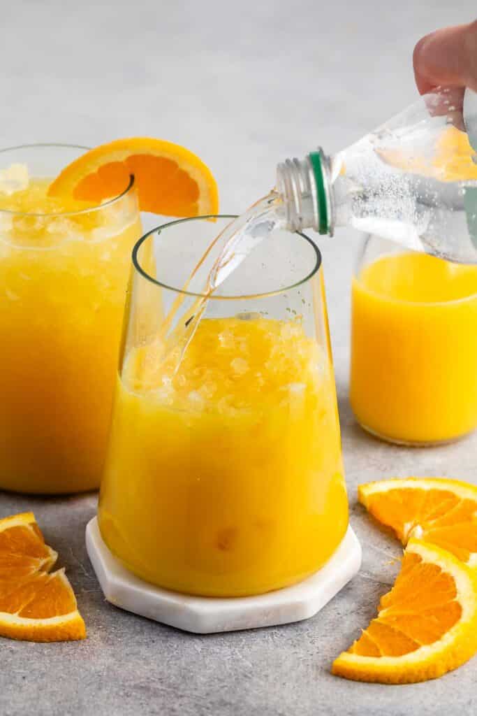 A refreshing orange drink is poured into a glass with ice, garnished with an orange slice. A hand pours clear soda from a bottle into the glass. There are more orange slices and a pitcher of juice in the background.