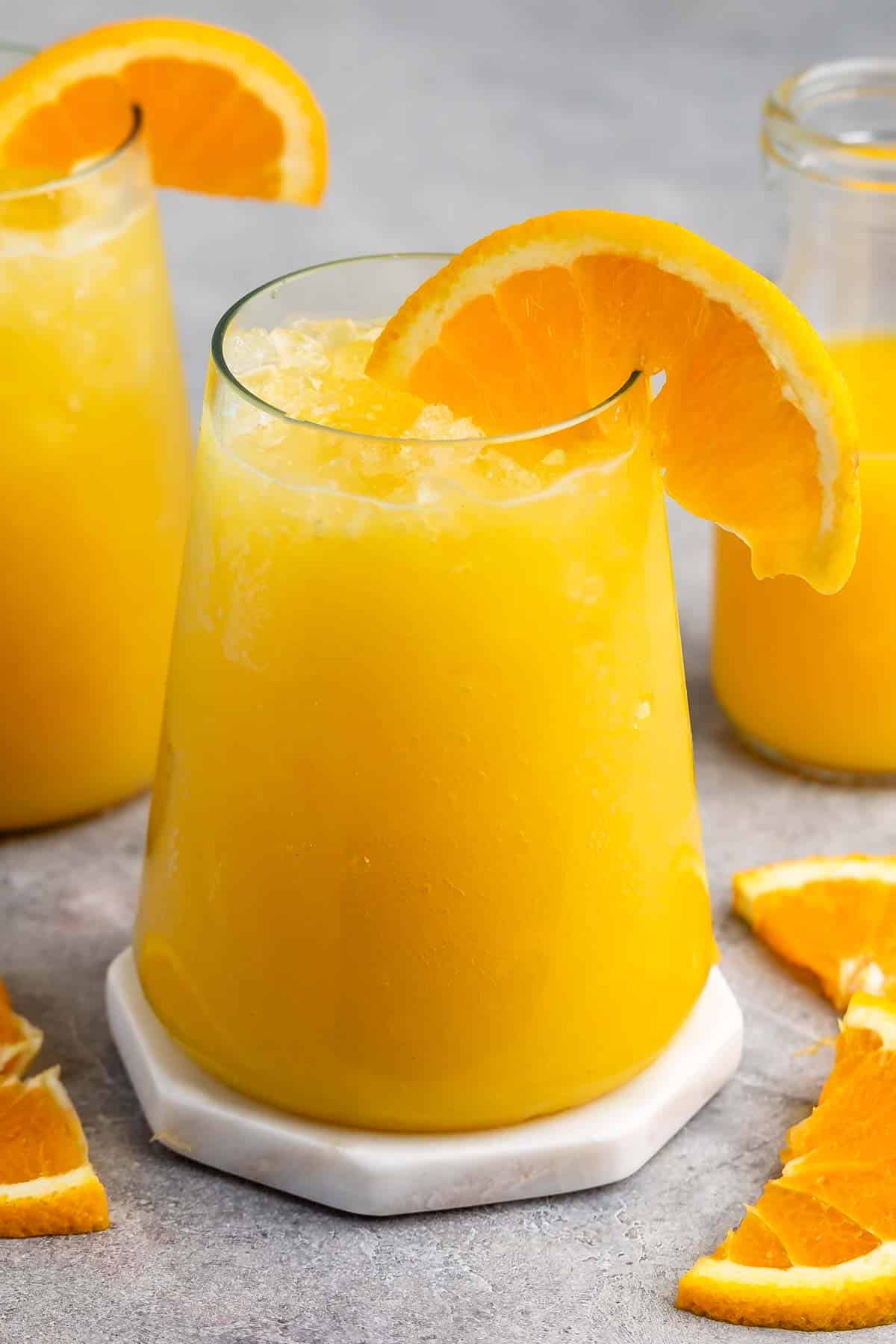 A glass of orange juice with crushed ice sits on a marble coaster. It is garnished with a slice of orange on the rim. Behind it, another glass and a bottle of orange juice are visible, surrounded by orange slices on a gray surface.