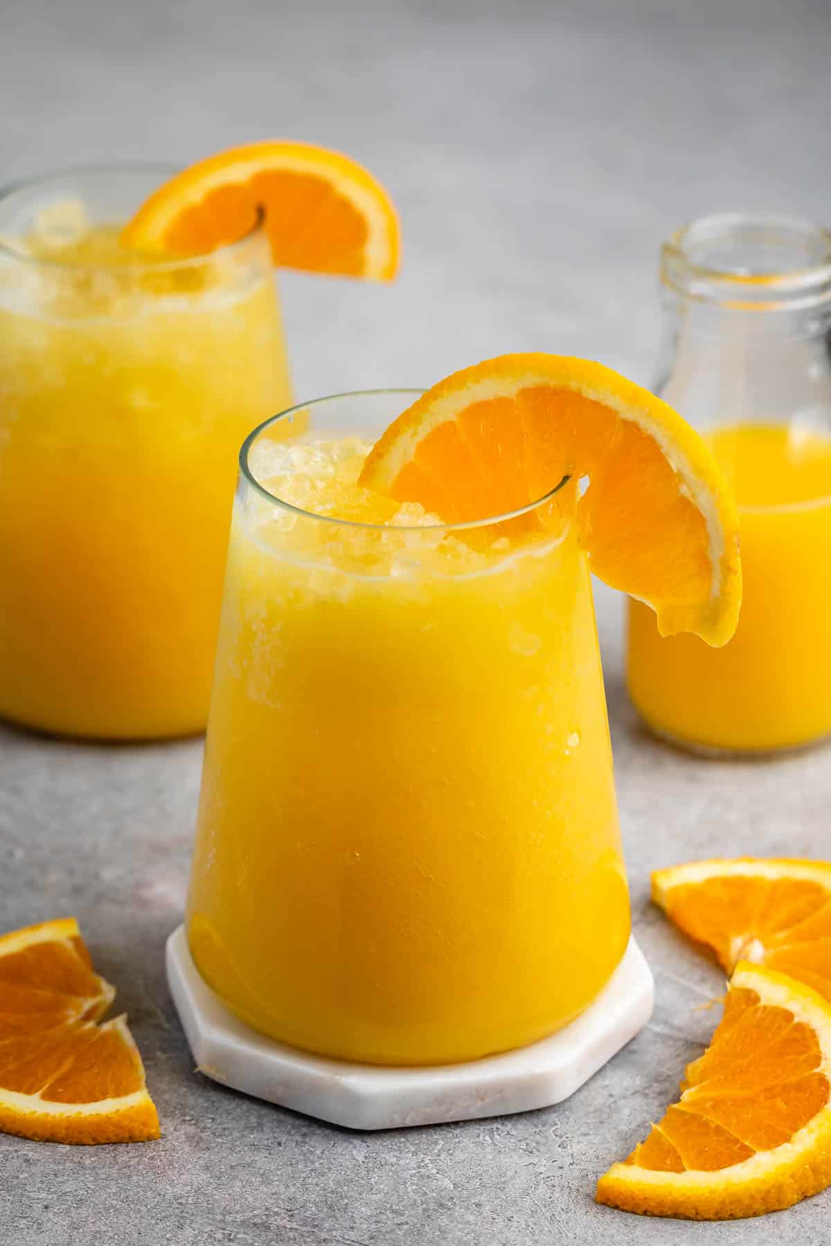 A refreshing orange smoothie in a glass, garnished with an orange slice. Additional orange wedges are scattered around on a gray surface. A small bottle of orange juice is visible in the background.