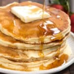 A stack of pancakes topped with a pat of butter, drizzled with syrup. Fresh strawberries and blueberries are blurred in the background. A fork is placed in front of the plate on a wooden table.