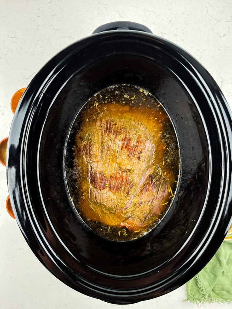 Top view of a slow cooker containing a cooked pot roast submerged in a flavorful broth. The roast is seasoned and surrounded by juices, with a light layer of seasoning visible on the surface. A green cloth and orange dishes are partially visible nearby.