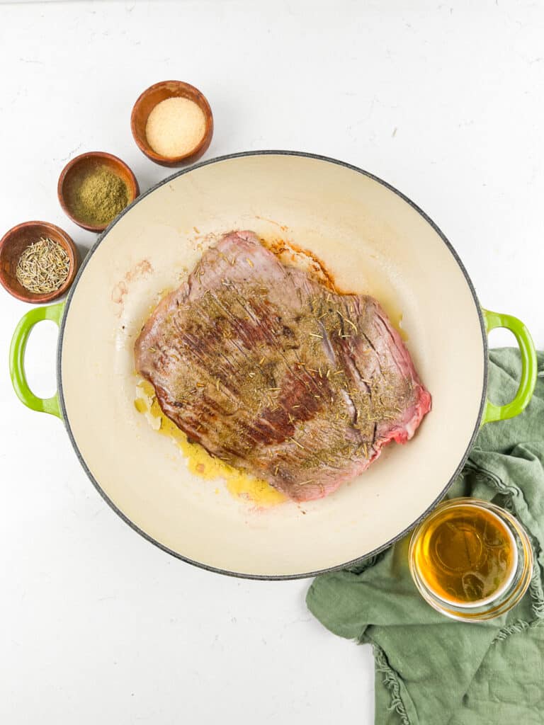 A large piece of seasoned beef is searing in a green-handled Dutch oven. Surrounding the pot are small bowls with various spices and a green cloth. A cup of liquid, possibly broth, is nearby on the white surface.