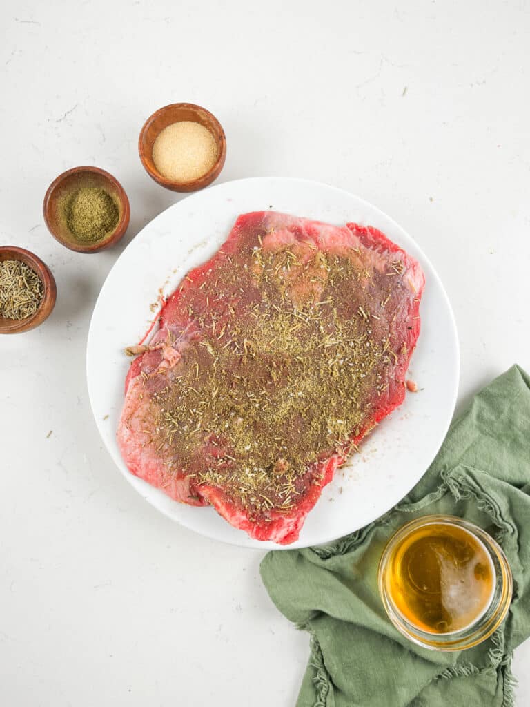 A plate of raw meat seasoned with herbs is on a white surface. Surrounding it are small bowls of spices and a small bowl of oil on a green cloth.