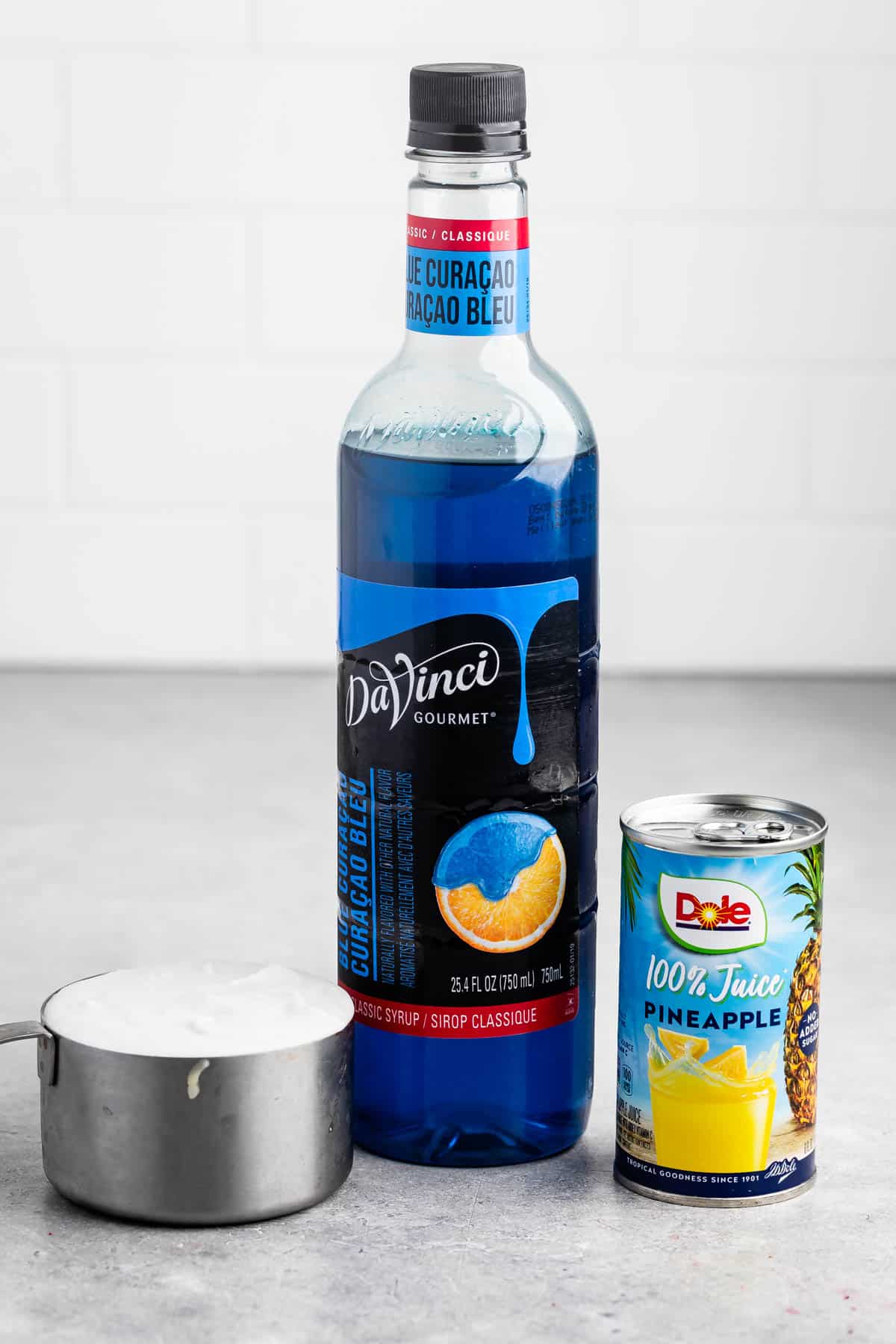 A bottle of DaVinci Gourmet Blue Curacao syrup stands next to a can of Dole 100% pineapple juice. In the foreground, theres a measuring cup filled with white liquid. The background features a white tile wall and a gray countertop.