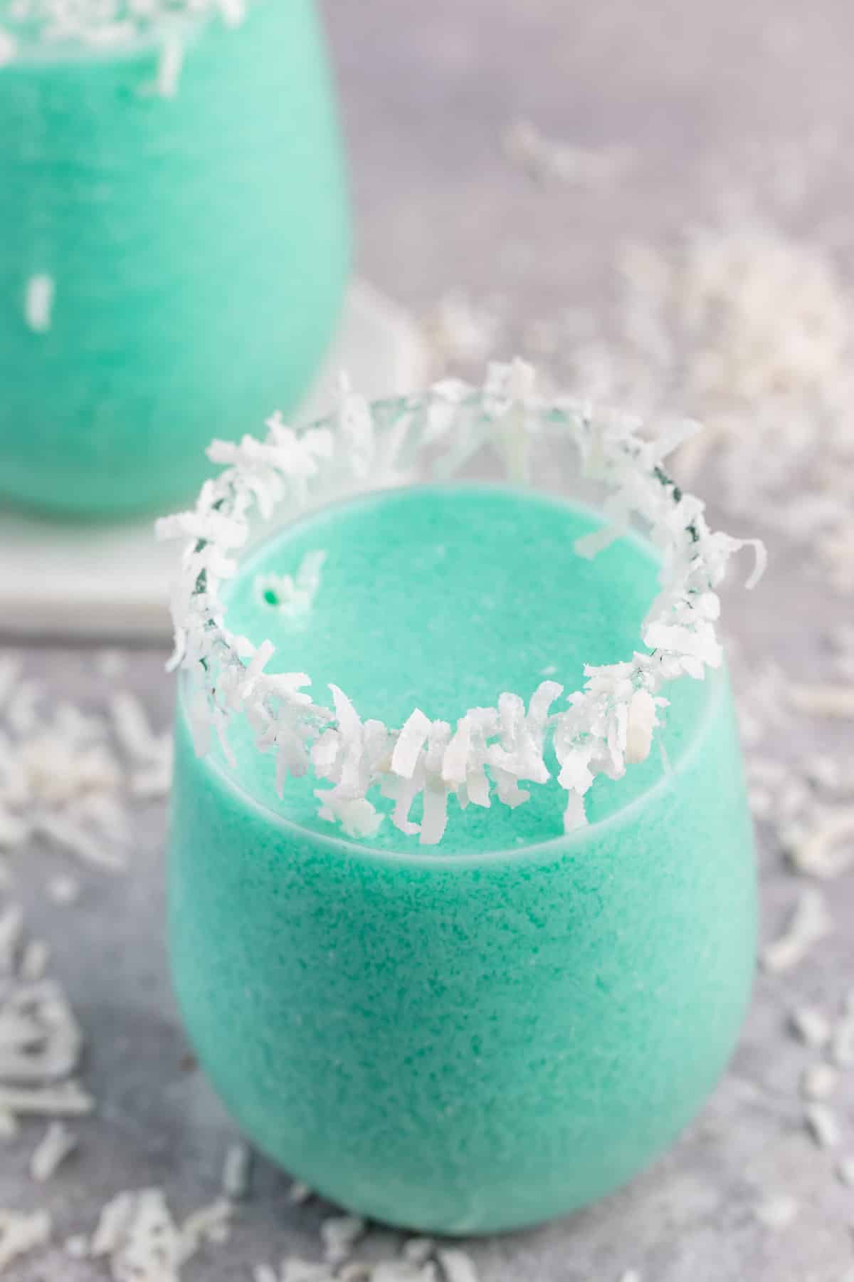 A vibrant blue-green tropical cocktail in a glass with a rim decorated with shredded coconut. The creamy drink sits on a gray surface scattered with more coconut flakes.