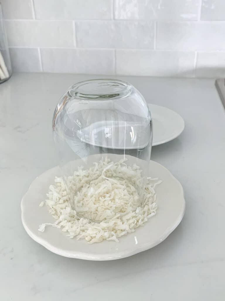 A glass placed upside down on a small plate filled with shredded coconut, set on a white countertop with a white tiled backsplash.