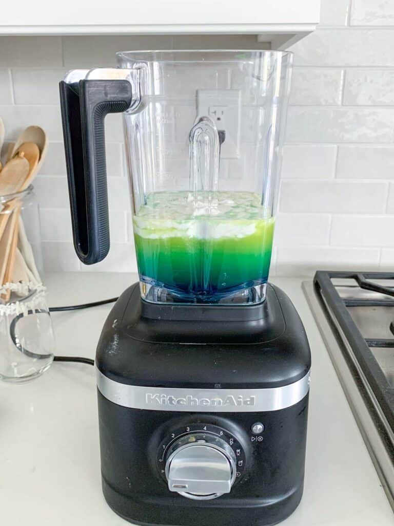 A black KitchenAid blender on a kitchen counter, filled with a green liquid. Wooden utensils in a jar and a stove are visible in the background.