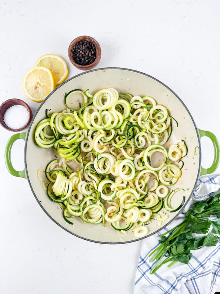 A large green pan filled with spiralized zucchini noodles on a white surface. Nearby are lemon halves, a bowl of black pepper, a bowl of salt, and fresh parsley on a striped cloth.