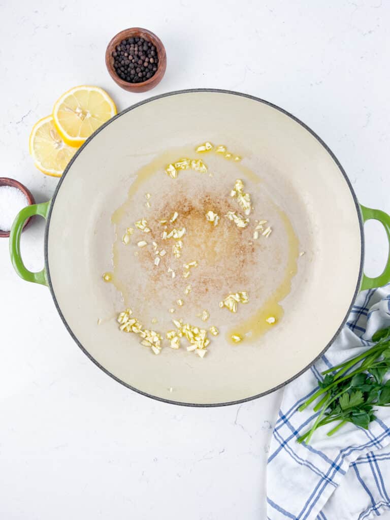 A large green-handled pot with olive oil and minced garlic on a white stove. Beside the pot are lemon slices, a bowl of peppercorns, salt, and fresh parsley on a checkered cloth.