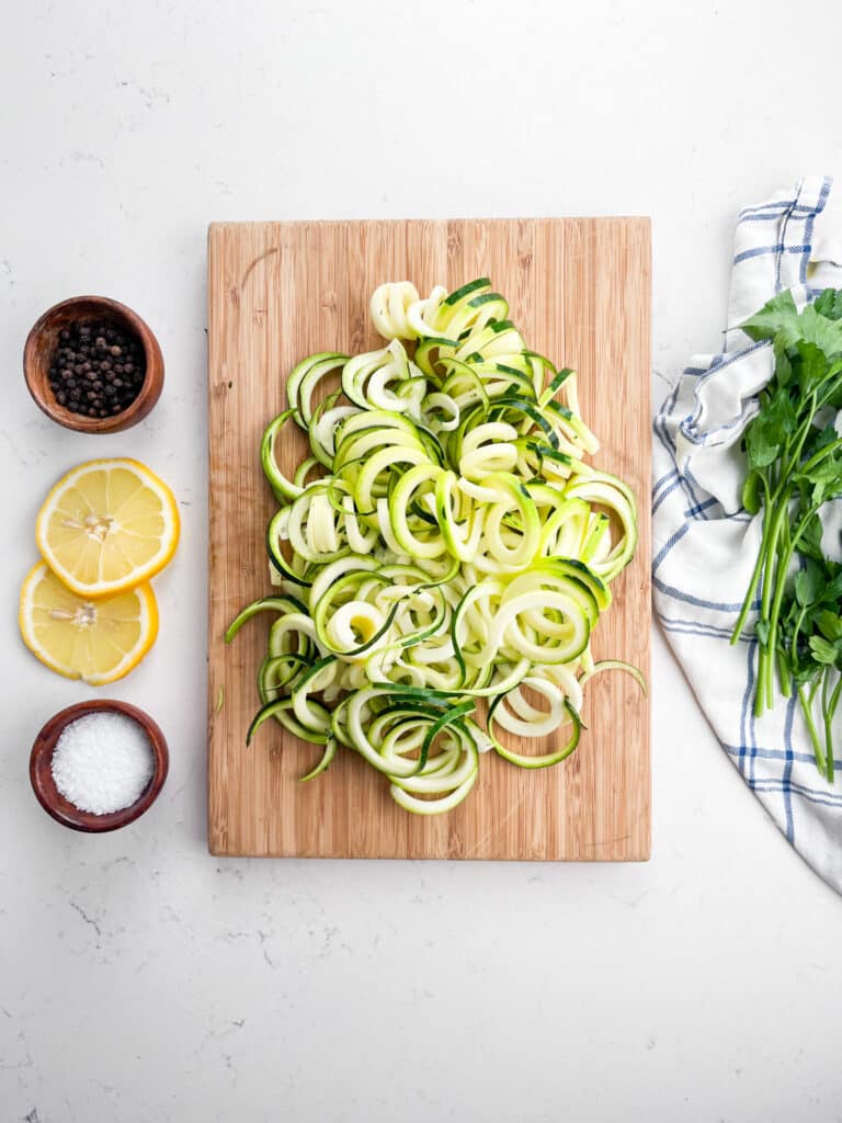 A wooden cutting board with spiralized zucchini sits on a white countertop. Beside it are a towel, fresh parsley, two lemon halves, a bowl of salt, and a bowl of peppercorns.