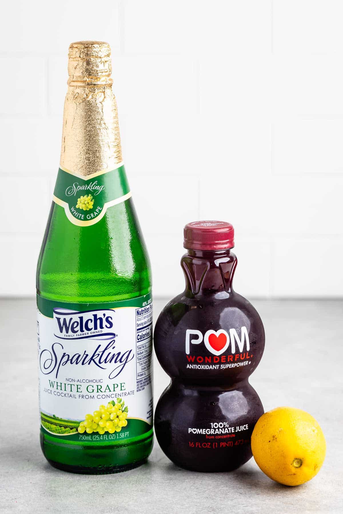 A bottle of Welchs Sparkling White Grape juice stands next to a bottle of POM Wonderful pomegranate juice. A lemon rests in front of them, all placed on a gray countertop with a white tiled wall in the background.